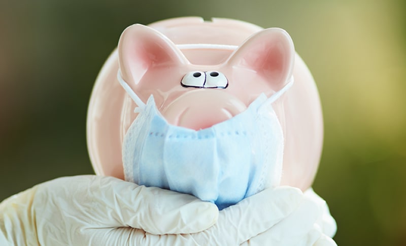 Ein Sparschwein mit medizinischer Maske wird von einer Hand mit Einmalhandschuhen gehalten.